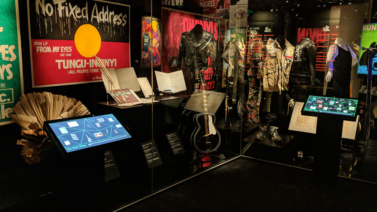 Australian music vault museum exhibition Wideshot of exhibition containing guitars, outfits and flags behind glass case.